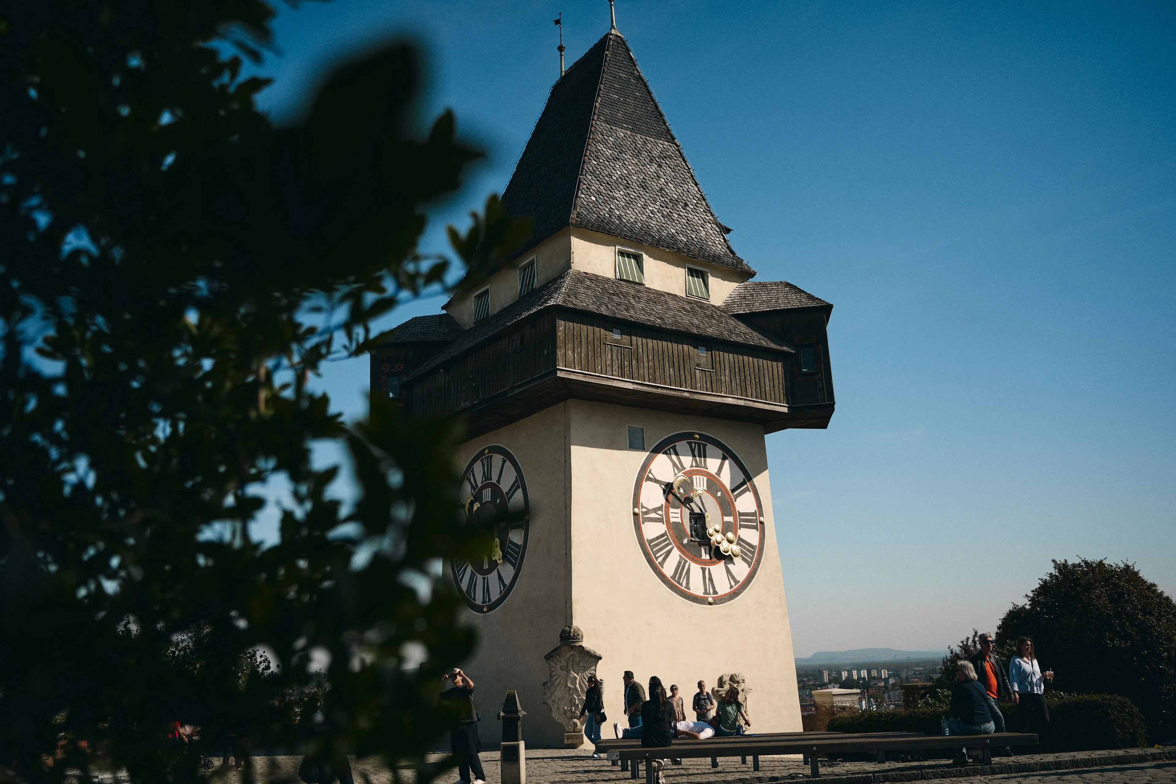 Der Uhrturm auf dem Schlossberg mit seinen großen Zifferblättern vor blauem Himmel, Besucher an seinem Fuß