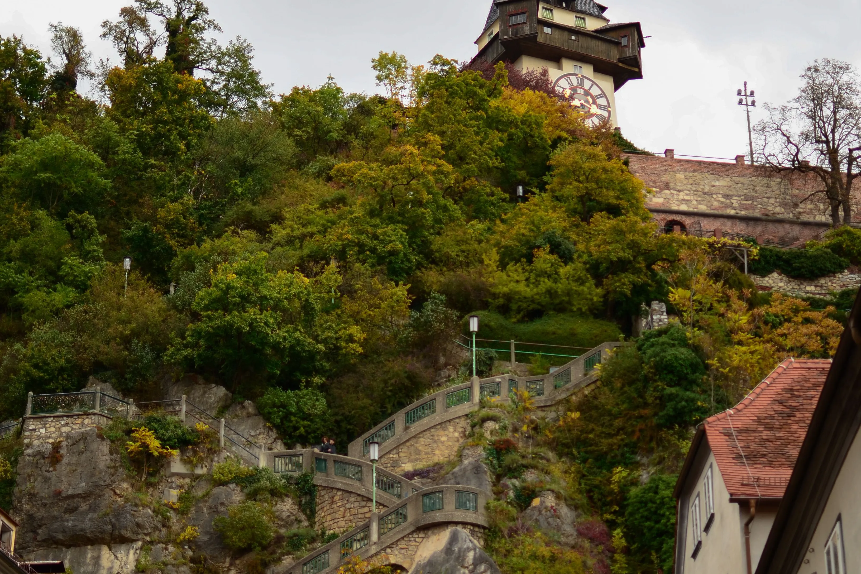 Steinerne Stiege in Kehren den Felsen des Schlossbergs hinauf, oben der Uhrturm zwischen Herbstbäumen