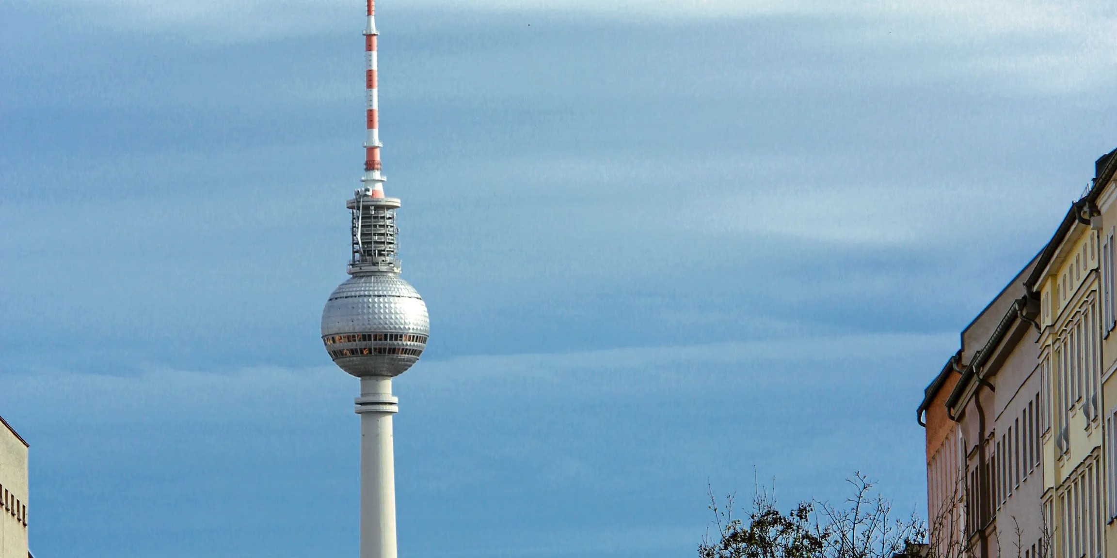 Street of old apartment buildings, trees and parked cars, with the TV tower at its end against a blue sky