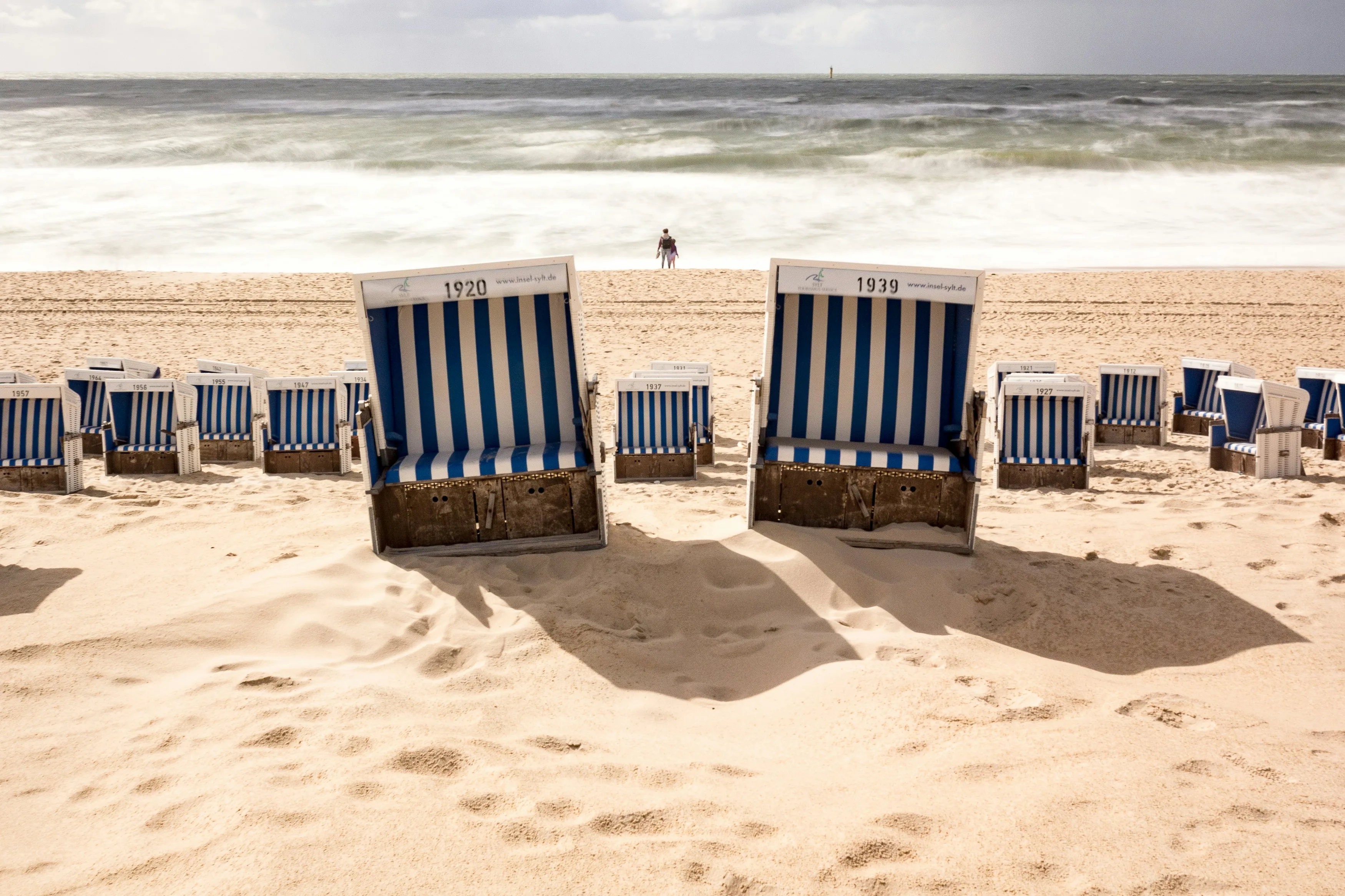 Blue and white beach chairs on a wide sandy beach in front of the foaming North Sea surf